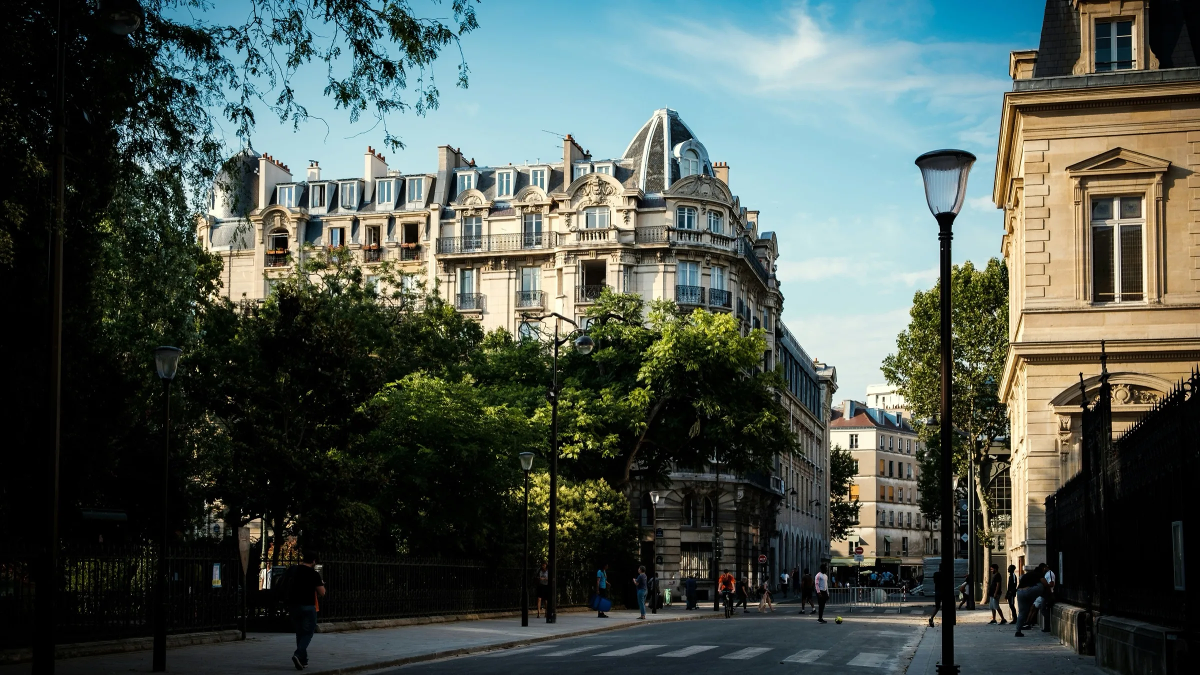Un immeuble d’angle à tourelle en dôme, sur une rue parisienne bordée d’arbres.