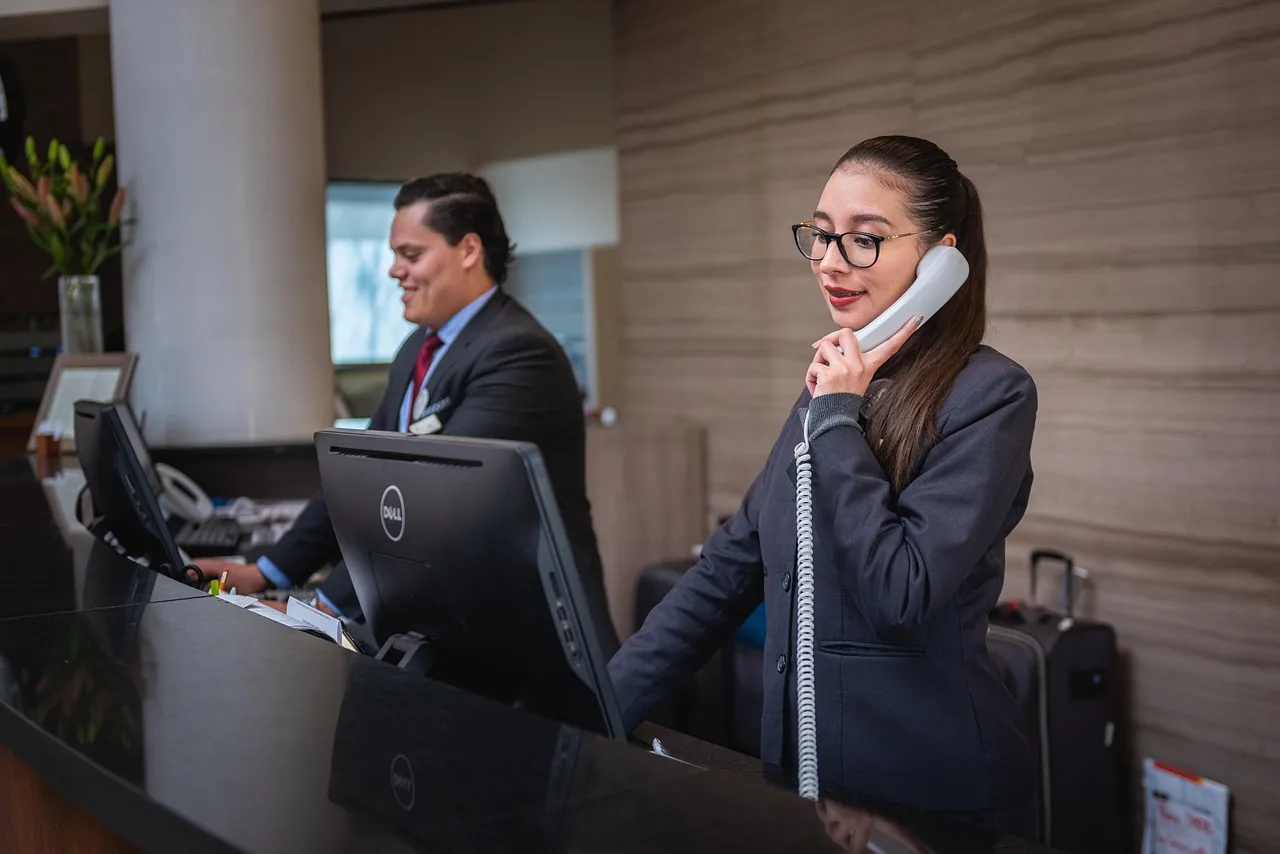 Une réceptionniste d'hôtel répondant au téléphone, avec la valise d'un client posée au comptoir.