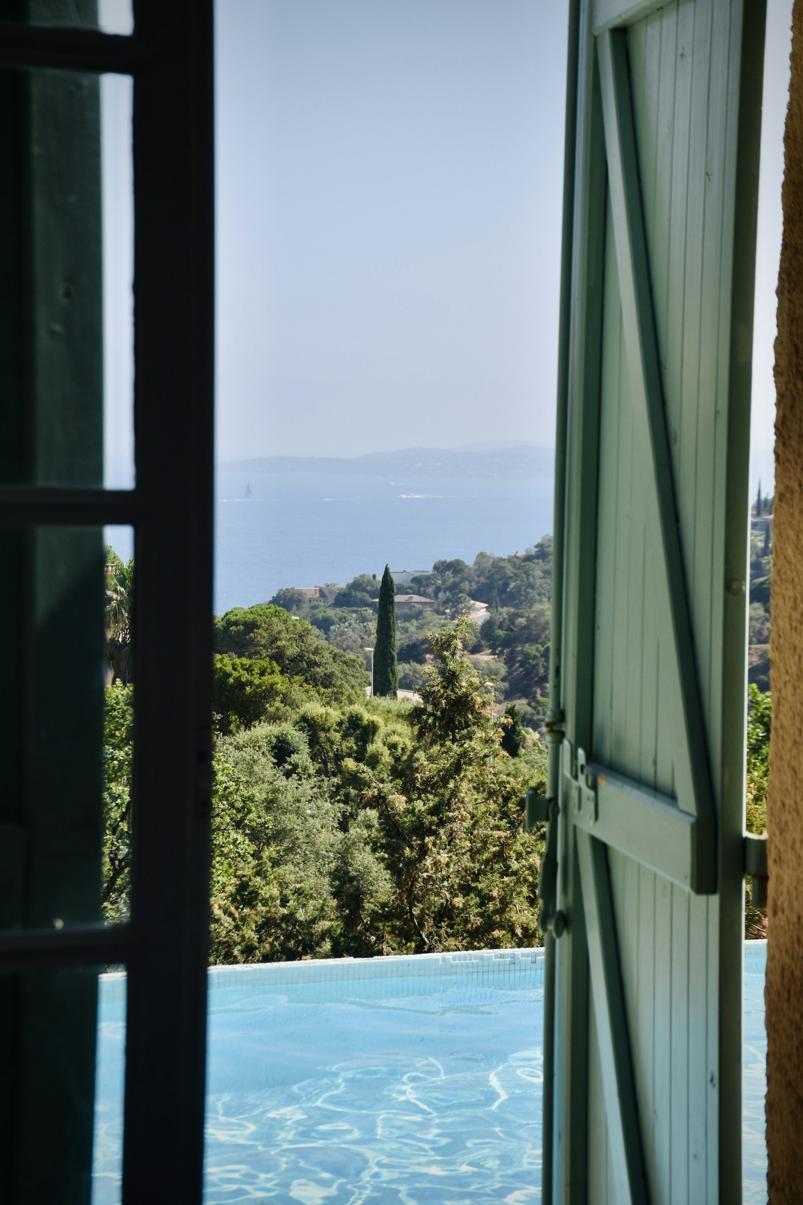 An open shuttered door framing a pool and sea view on the French Riviera coast.