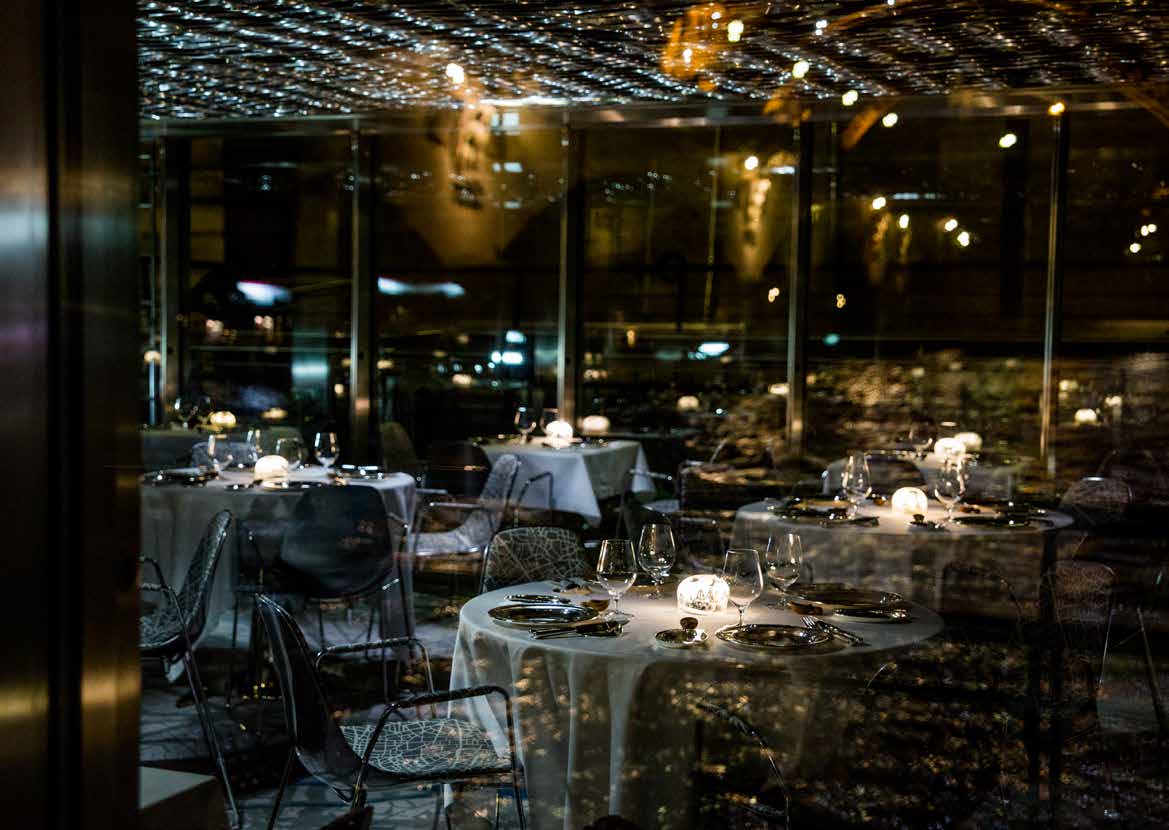 Des tables aux chandelles dans la salle vitrée de Ducasse sur Seine, la nuit.
