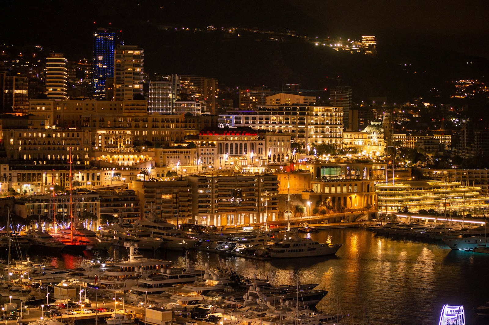 Monaco's harbour and hillside skyline lit up at night, yachts moored along the quay.
